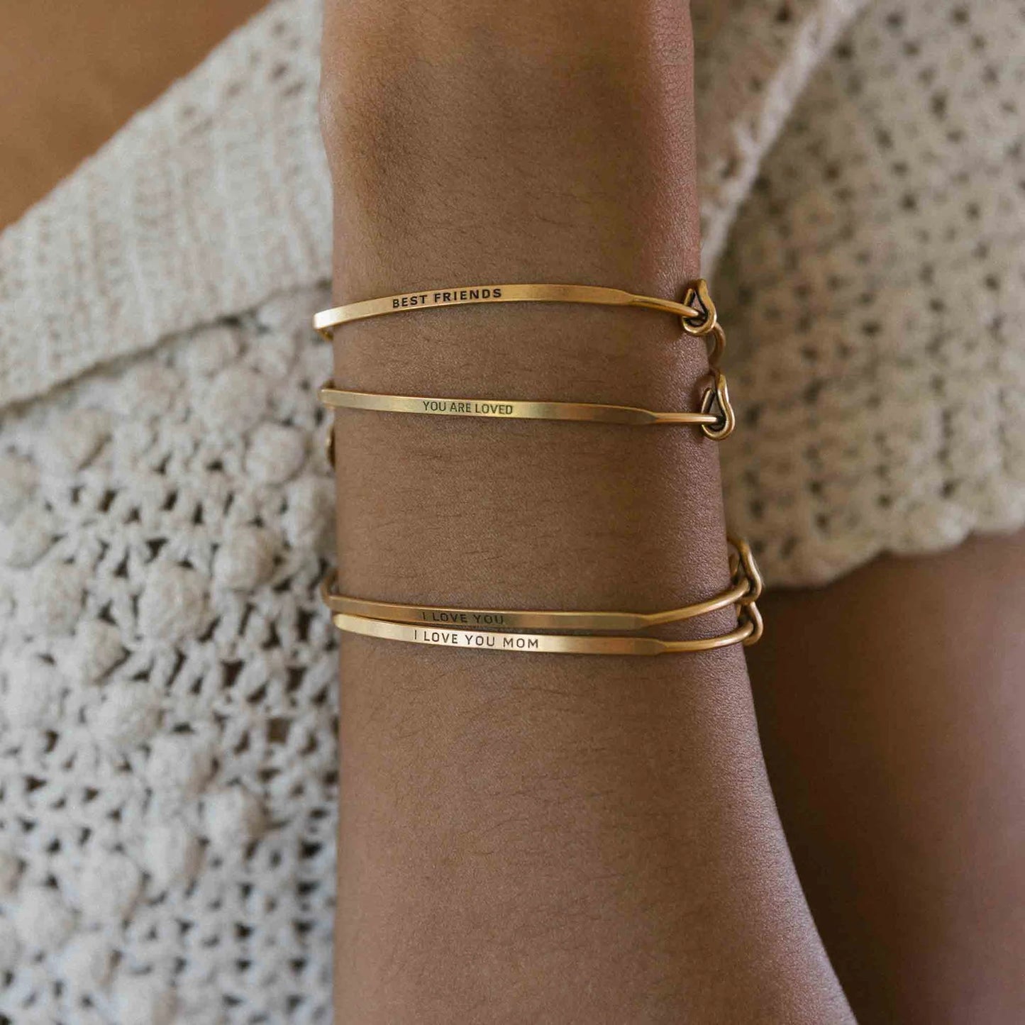 Gold bracelets with engraved messages on a wrist against a textured fabric background
