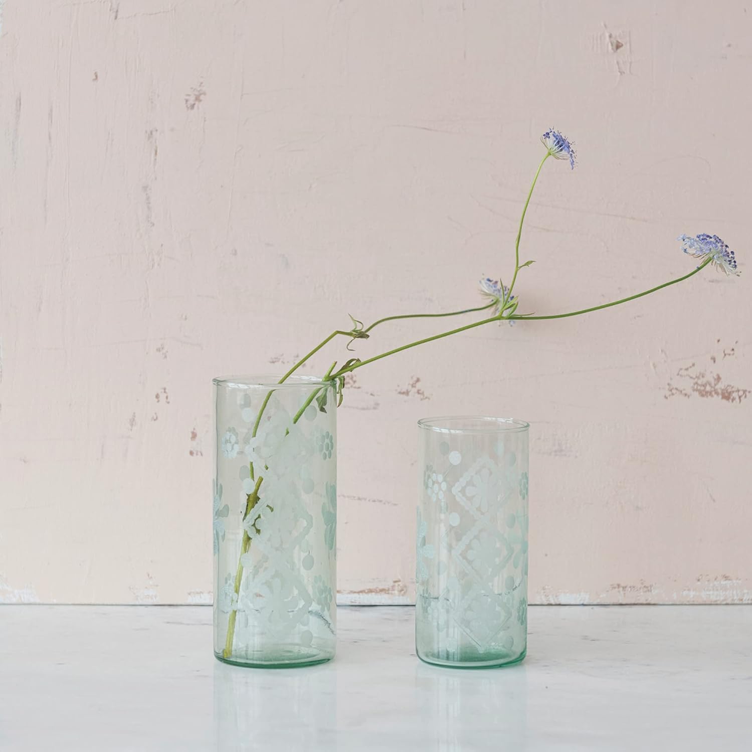 Two textured glass vases with a single stem of flowers against a light pink background