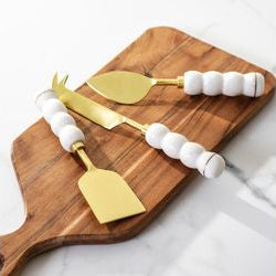 Three gold cheese knives with white marble handles on a wooden cutting board.