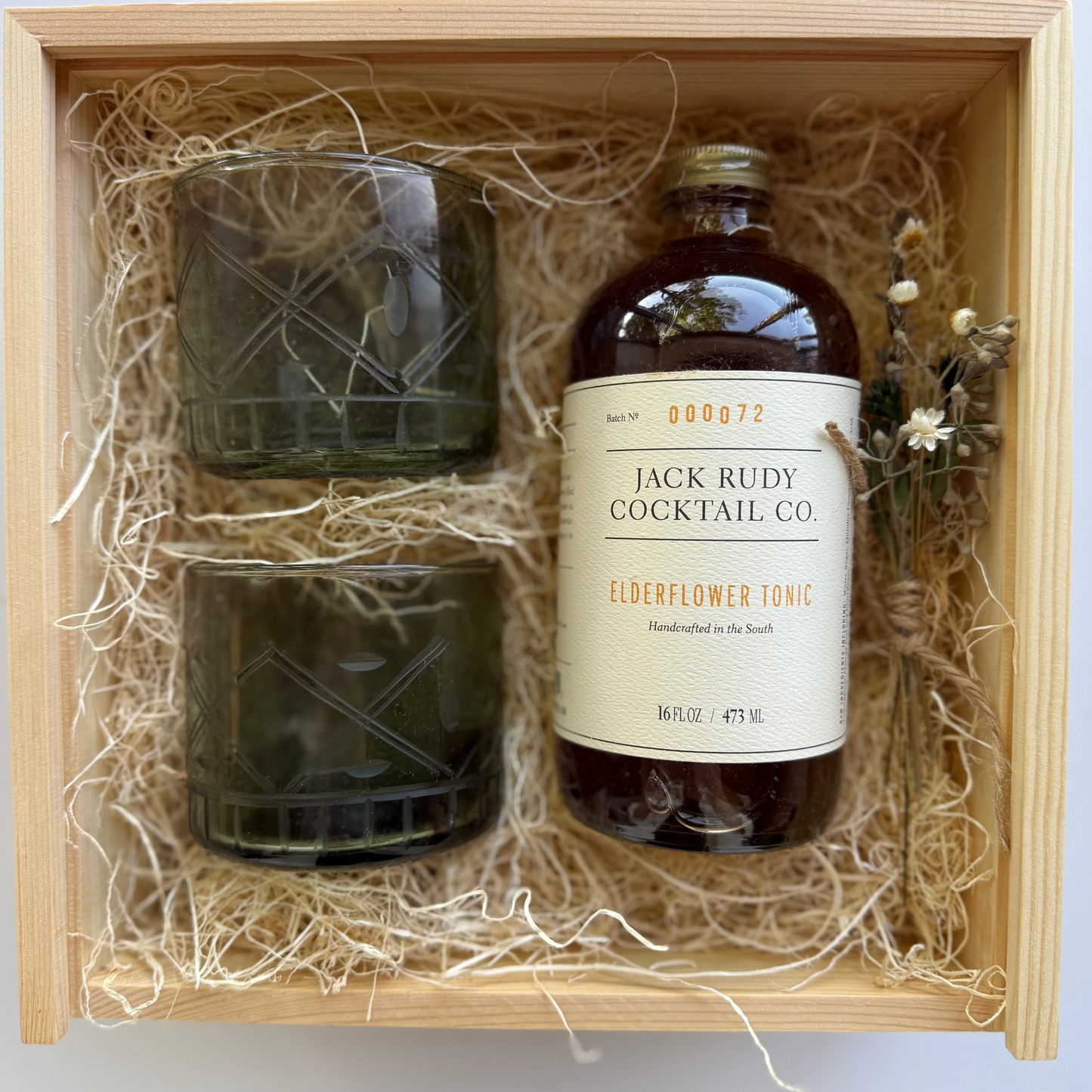 Bottle of Jack Rudy Elderflower Tonic and two glasses in a wooden box with straw lining.