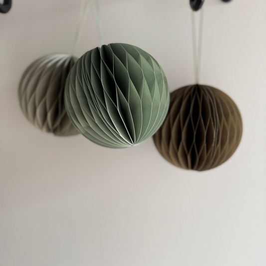 Three paper honeycomb balls in green, gray, and brown hanging against a white wall.
